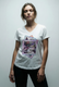 A woman with light brown hair in a loose updo wears The VIV Tee By You by SUZI ROHER, a customizable V-neck with a colorful modern lounge graphic, standing against a plain light background and facing the camera neutrally.