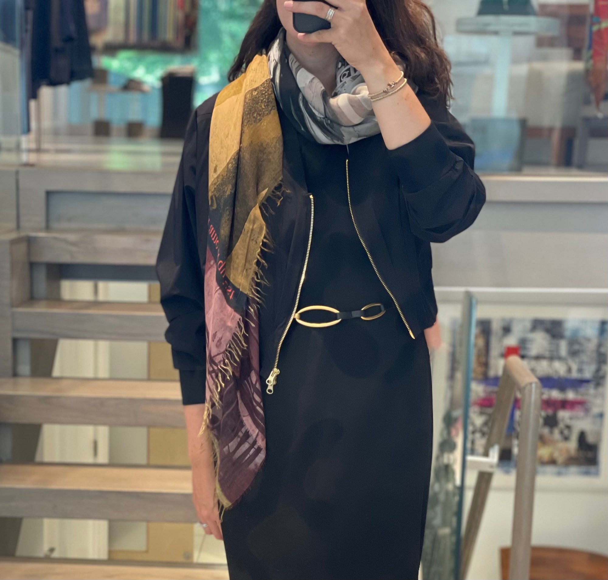 A women wearing a scarf with a black and white center and warm gold and Burgundy tones around the edge that falls down her black dress. 