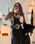 A chic person with wavy brown hair takes a mirror selfie in a modern, stylish room, wearing the Moda Florentina by SUZI ROHER: a sheer black top, printed scarf, wide belt with gold buckle, and holding a black furry coat.