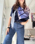 A woman stands indoors in a black top, floral wide-leg pants, and the SUZI ROHER Vogue Covers V.02 scarf, channeling classic magazine style with one hand on her hip against a brightly modern, softly blurred background.