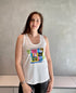 A woman with long brown hair wears The Lola, a white sleeveless top with colorful graphic prints from SUZI ROHER, paired with black pants, standing confidently by a gray wall and white cabinets.