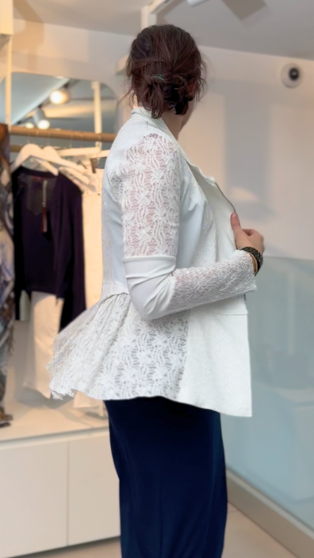 A woman with dark hair in a low bun wears the HIGH Affair Jacket, a white lace blazer with a peplum back, paired with a navy skirt, standing sideways in a modern clothing store with racks of clothes behind her.