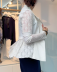 A woman with dark hair in a low bun wears the HIGH Affair Jacket, a white lace blazer with a peplum back, paired with a navy skirt, standing sideways in a modern clothing store with racks of clothes behind her.