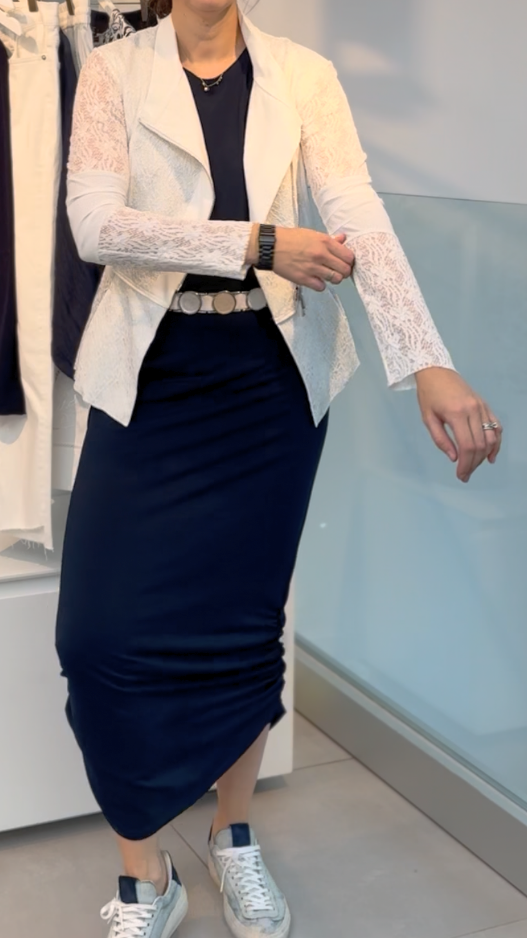 Indoors, a person adjusts the sleeve of their HIGH Affair Jacket with lace sleeves, styled with a navy maxi skirt, black top, white belt, and white sneakers.