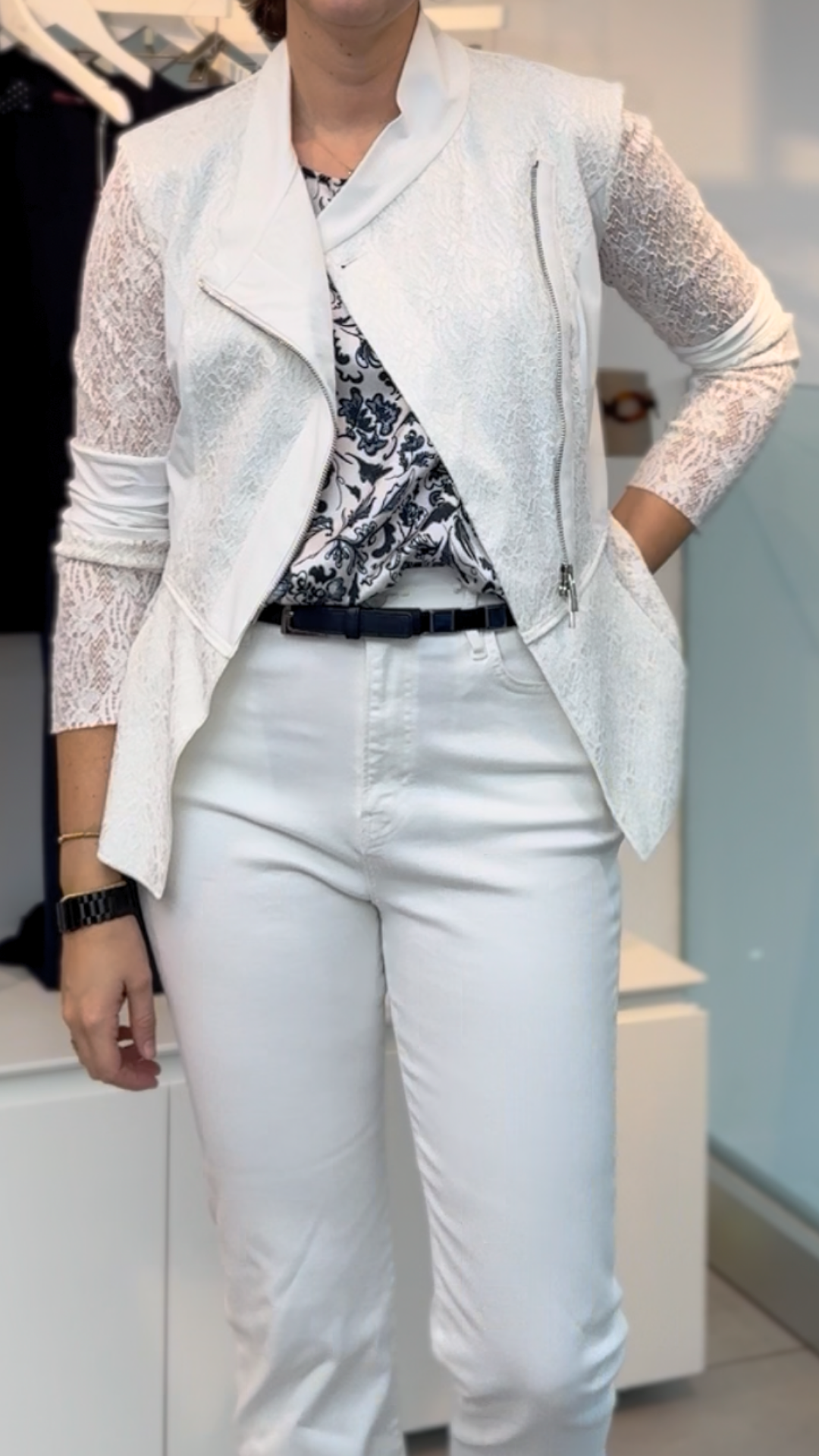 A woman stands indoors in sophisticated style, wearing white pants, a black-and-white patterned blouse, and the HIGH Affair Jacket with an asymmetrical zipper. She completes her look with a black watch and belt.