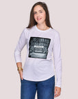 A woman with long brown hair wears SUZI ROHER’s The Catherine—a white long-sleeve shirt featuring a black-and-white APPLE storefront photo—standing against a light gray background, softly smiling in this chic, understated look.