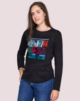 A woman with long brown hair stands against a plain light background, wearing SUZI ROHER's The Catherine—a 100% cotton black crew neck tee with a colorful graphic and the word "captcha"—paired with blue jeans.