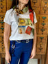 A woman stands before vintage wooden drawers, wearing SUZI ROHER's The Karlie white T-shirt featuring vintage Vogue covers, paired with a colorful scarf, blue pants, and a statement belt. Her head is partially out of frame.