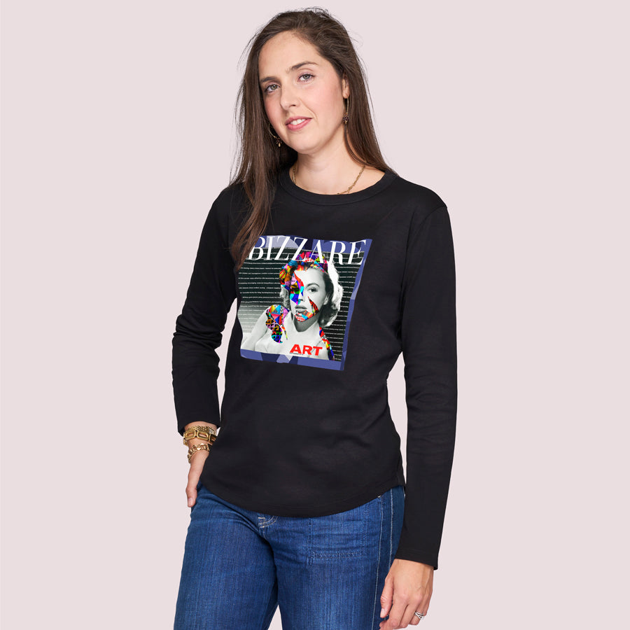 A woman with long brown hair wears The Catherine by SUZI ROHER, a 100% cotton black crew neck long-sleeve shirt featuring a colorful artistic graphic and the word “BIZZARE” on the front. She smiles slightly, one hand in her pocket.