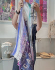 A women opening and putting on a scarf to match her outfit. The scarf is pink, purple and blue with a graphic of Café interior with coffee cups, saucers, and a pen on a table, featuring the name 'L'Escargot'