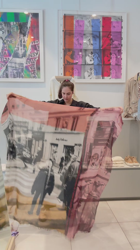 A women showing the Color of Love scarf, featuring  multicoloured abstract pattern—burgundy, gold, pink, black, and cream with frayed edges and a soft texture. She then wraps it around her neck.