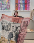 A women showing the Color of Love scarf, featuring  multicoloured abstract pattern—burgundy, gold, pink, black, and cream with frayed edges and a soft texture. She then wraps it around her neck.