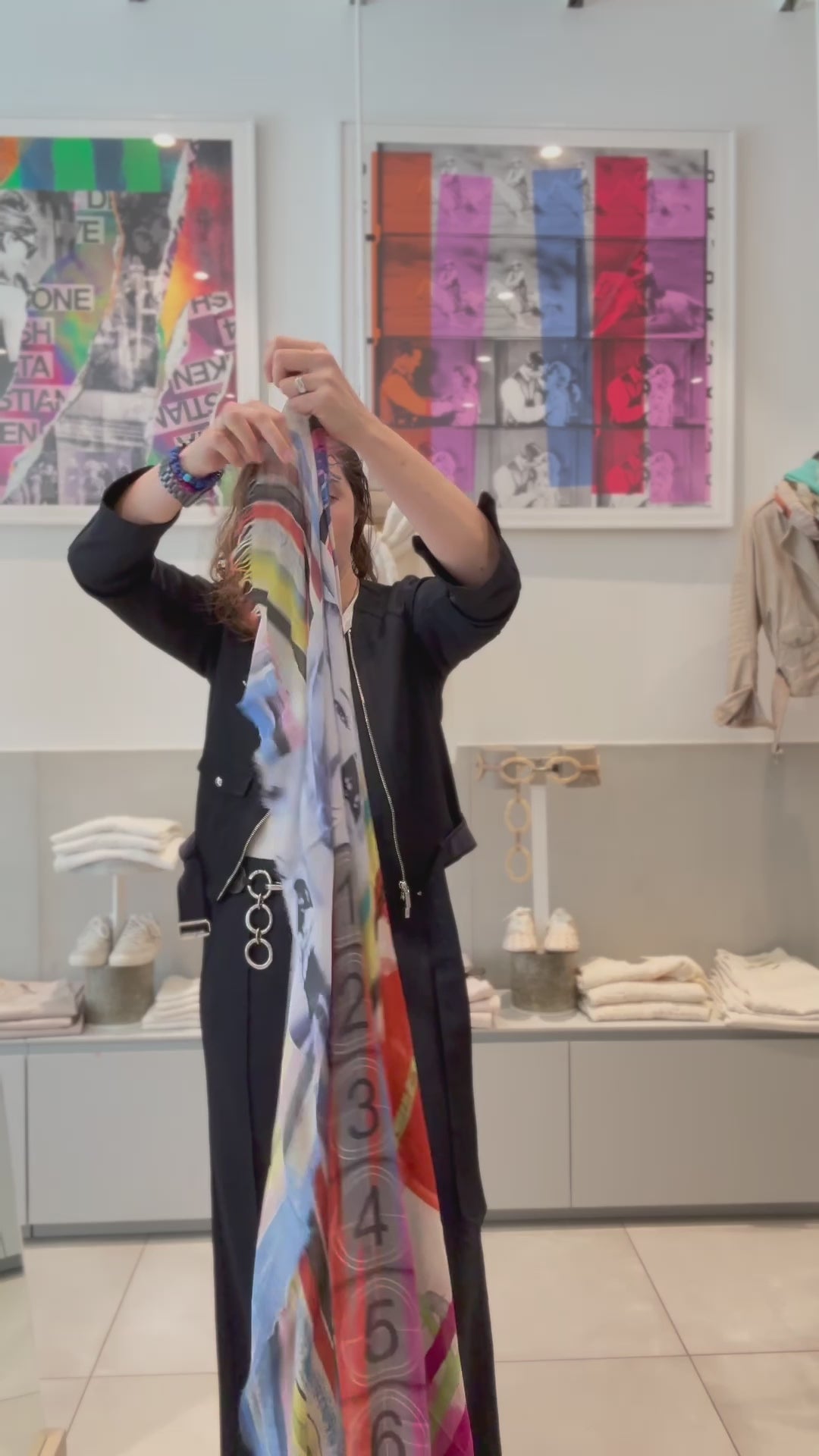 A video of a women putting on a the SUZI ROHER just a second scarf. The SUZI ROHER Just a Sec scarf features frayed edges and a vibrant, multicolored abstract design, recalling old Hollywood glamour with its swirling stripes—a timeless style inspired by icons like Audrey Hepburn and Marilyn Monroe.