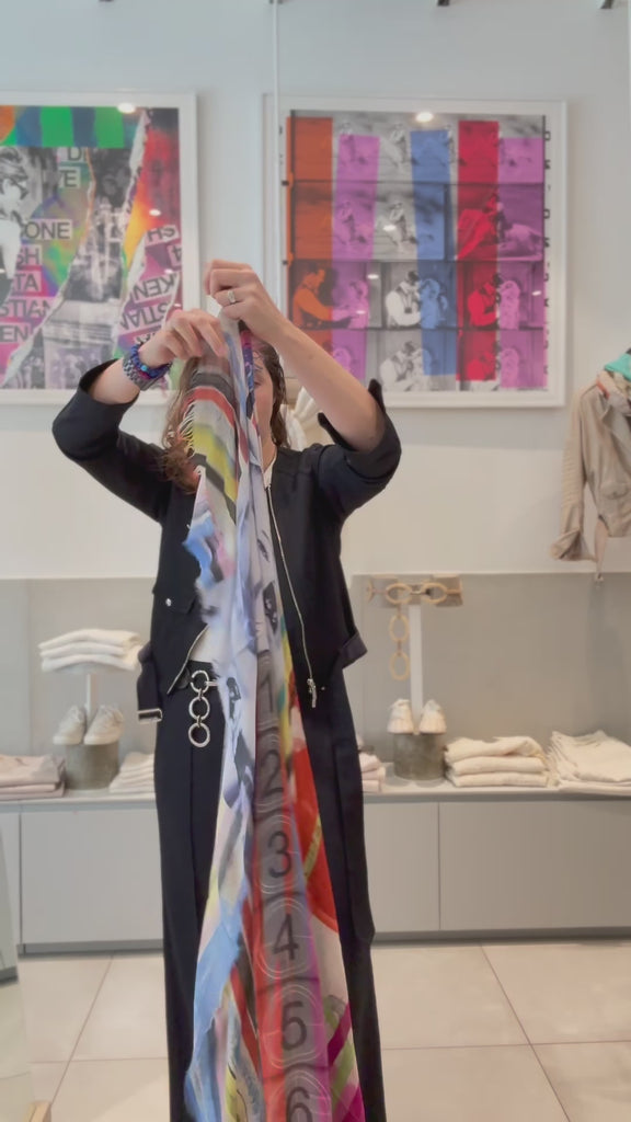A video of a women putting on a the SUZI ROHER just a second scarf. The SUZI ROHER Just a Sec scarf features frayed edges and a vibrant, multicolored abstract design, recalling old Hollywood glamour with its swirling stripes—a timeless style inspired by icons like Audrey Hepburn and Marilyn Monroe.