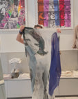 A square scarf, being shown open, then wrapped onto a women's neck. The scarf has original artwork of, a black and white photo of a Jackie-O with a different blue square overlay on the sides. 