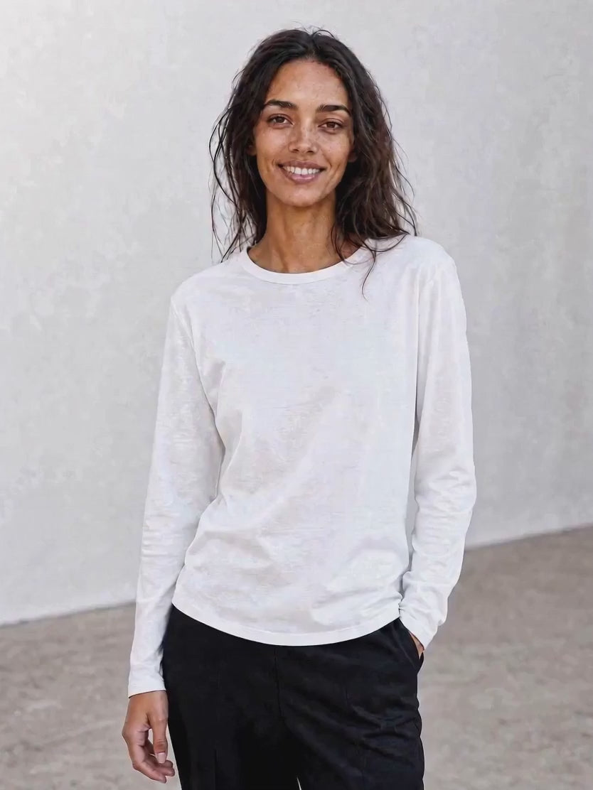 A woman with long dark hair, dressed in The CATHERINE Tee By You by SUZI ROHER—a luxury crew neck white long-sleeve shirt—paired with light pants, stands against a plain light background, smiling with her hands in her pockets.