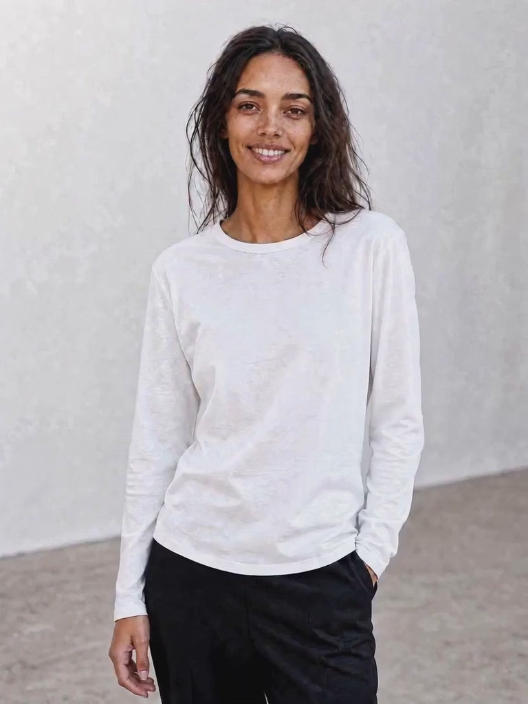A woman with long dark hair, dressed in The CATHERINE Tee By You by SUZI ROHER—a luxury crew neck white long-sleeve shirt—paired with light pants, stands against a plain light background, smiling with her hands in her pockets.