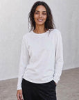 A woman with long dark hair, dressed in The CATHERINE Tee By You by SUZI ROHER—a luxury crew neck white long-sleeve shirt—paired with light pants, stands against a plain light background, smiling with her hands in her pockets.