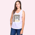 A woman with long brown hair smiles at the camera in "The Lola" by SUZI ROHER, a sleeveless white graphic top paired with blue jeans, standing against a light pink background.