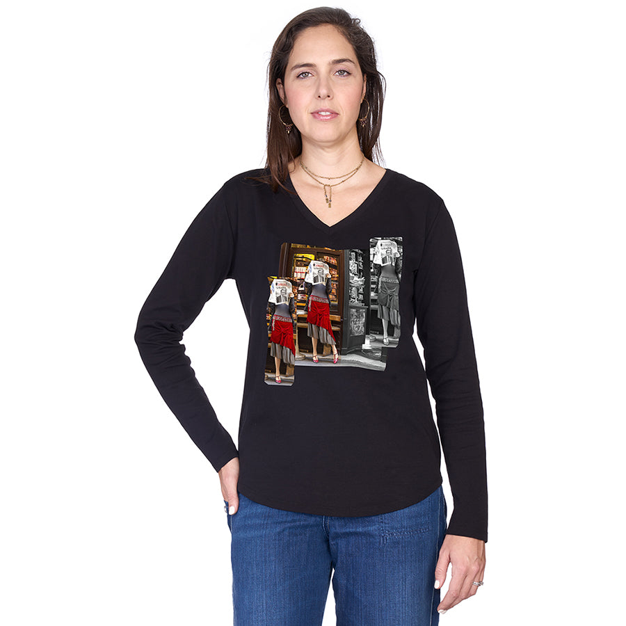 A woman with shoulder-length brown hair models SUZI ROHER’s The Victoria—a black 100% cotton long-sleeve tee featuring a vibrant graphic of three vintage-dressed women by a photo booth—paired with blue jeans and one hand in her pocket.