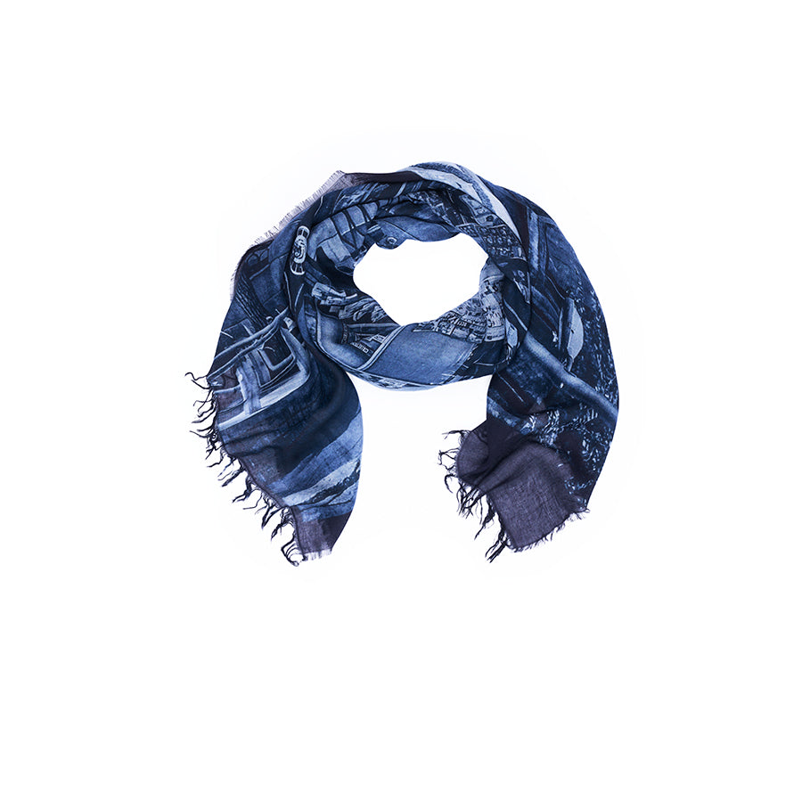 A SUZI ROHER scarf, named “Lily, Vogue Italia B&W,” featuring a subtle, digitally printed pattern and frayed edges in dark blue tones, is loosely coiled in a circular shape on a white background.