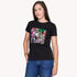 A woman with long brown hair models SUZI ROHER's "The Niki"—a black graphic T-shirt with a vibrant Breakfast at Tiffany's–inspired collage—paired with blue jeans, standing against a plain white background, one hand in her pocket.