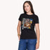 A woman with long brown hair models The Niki slim fit black crewneck tee by SUZI ROHER, crafted from 100% cotton with a vibrant artistic collage on the front, paired with blue jeans against a plain light background.