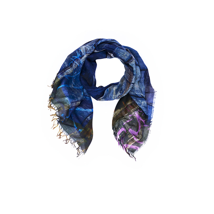 A loosely folded indigo scarf called “Lily, Against All Odds” from SUZI ROHER, featuring abstract patterns, frayed edges, and Italian-made digital printing, displayed on a white background.