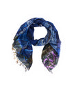 A loosely folded indigo scarf called “Lily, Against All Odds” from SUZI ROHER, featuring abstract patterns, frayed edges, and Italian-made digital printing, displayed on a white background.