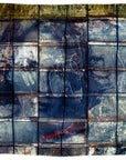 A striking scene reminiscent of a grid print on an Italian-made indigo scarf, the SUZI ROHER “Lily, Against All Odds” features a large, weathered blue metal grid adorned with graffiti and chalk markings and the words “Against All Odds.” faintly printed in red near the bottom center.