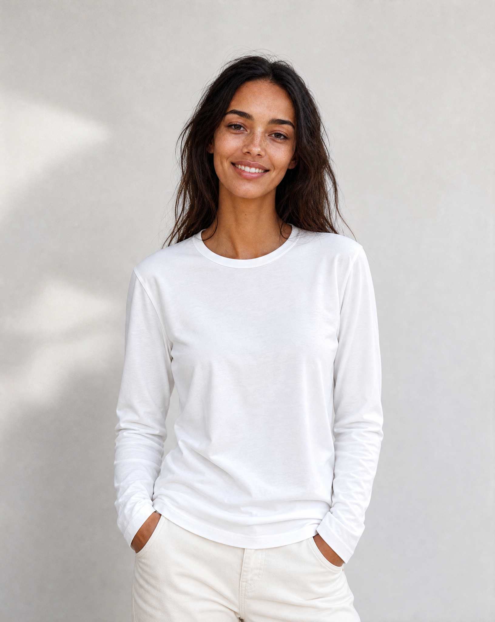 A woman with long dark hair, dressed in The CATHERINE Tee By You by SUZI ROHER—a luxury crew neck white long-sleeve shirt—paired with light pants, stands against a plain light background, smiling with her hands in her pockets.