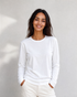 A woman with long dark hair, dressed in The CATHERINE Tee By You by SUZI ROHER—a luxury crew neck white long-sleeve shirt—paired with light pants, stands against a plain light background, smiling with her hands in her pockets.