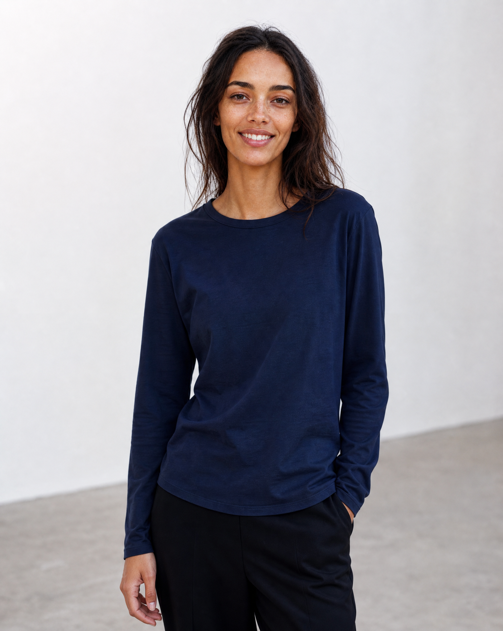 A woman with long dark hair, dressed in the SUZI ROHER “The CATHERINE Tee By You” navy blue customized long-sleeve tee and black pants, stands indoors against a light-colored wall, smiling at the camera.