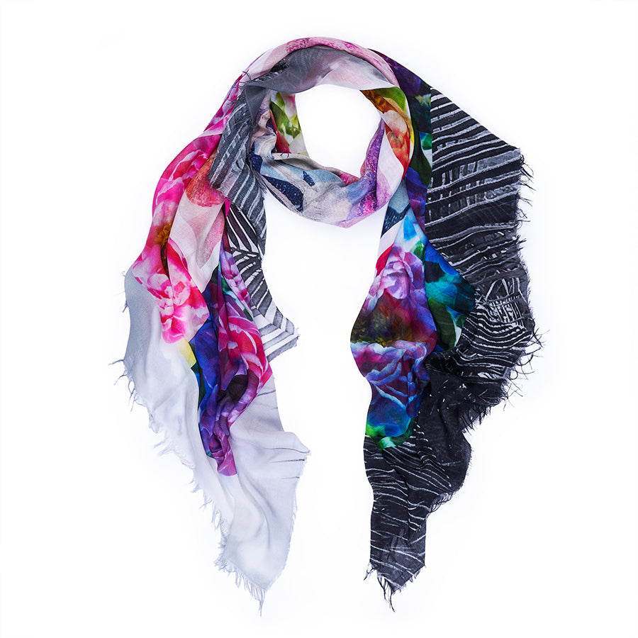 The SUZI ROHER Garden Party scarf is a lightweight, oversized Italian silk piece with fringed edges, featuring pink, purple, and blue florals on one side and black-and-white diagonal stripes on the reverse.