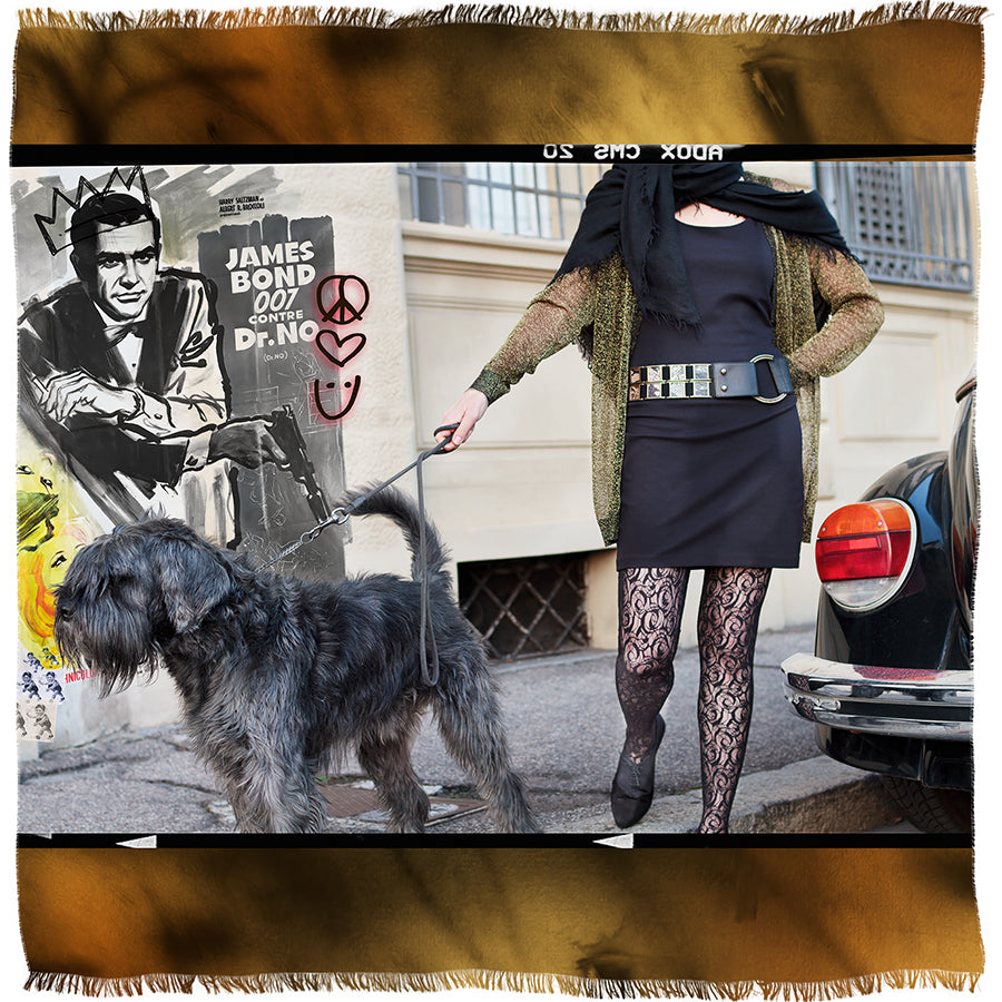 A woman in a black dress and patterned tights walks a large, shaggy black dog down a city street. Behind her is a mural featuring SUZI ROHER’s James Bond and other street art symbols.