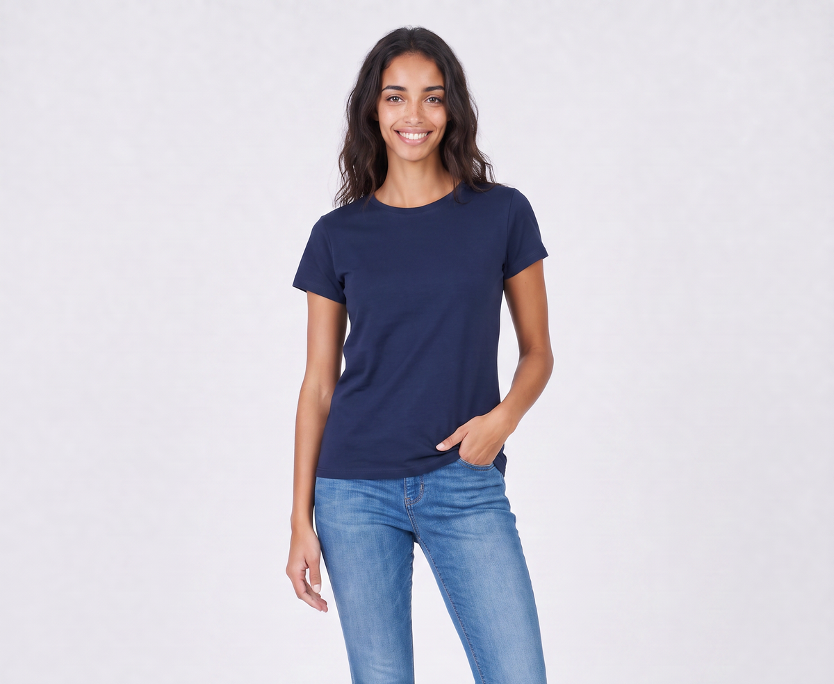 A woman with long, wavy dark hair smiles while standing against a light background. She wears The KARLIE Tee By You in navy blue by SUZI ROHER and blue jeans, with one hand in her pocket.