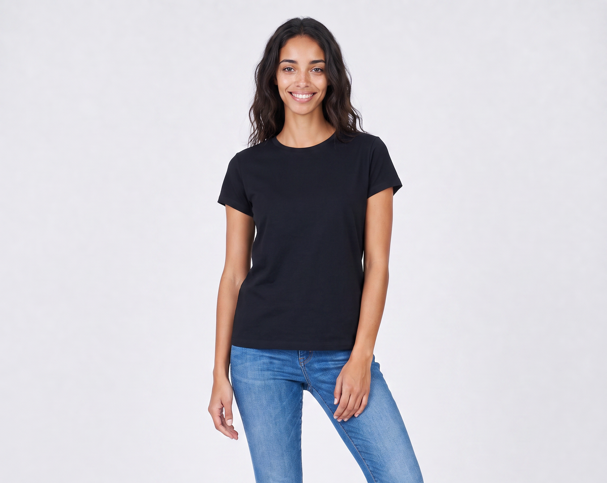 A woman with long dark hair smiles while standing against a light background. She is wearing The KARLIE Tee By You, a plain black customizable t-shirt by SUZI ROHER, and blue jeans.