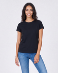 A woman with long dark hair smiles while standing against a light background. She is wearing The KARLIE Tee By You, a plain black customizable t-shirt by SUZI ROHER, and blue jeans.
