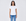 A woman with long dark hair stands smiling, wearing The KARLIE Tee By You customizable white t-shirt by SUZI ROHER and blue jeans against a light gray background.