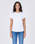 A woman with long dark hair stands smiling, wearing The KARLIE Tee By You customizable white t-shirt by SUZI ROHER and blue jeans against a light gray background.