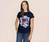 A woman with long brown hair models The KARLIE Tee By You by SUZI ROHER, a navy customizable t-shirt with a colorful framed photo graphic, standing with one hand in her jeans pocket against a plain light background.