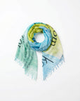 The SUZI ROHER Lily, Yugen scarf features a lightweight design with frayed edges and a gradient of light green, yellow, and blue, overlaid by black text in various directions. Presented elegantly coiled against a white background.
