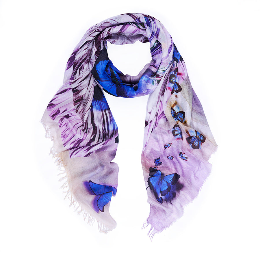 The SUZI ROHER Social Butterfly scarf features blue and purple butterfly prints on light purple fabric with fringed edges, displayed in a loose loop against a white background.