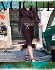 A woman in a brown dress and sandals walks past a green car and vintage posters on a weathered street, her image transformed into original artwork with an elegant Italian finish by SUZI ROHER, featured on the cover of Spanish Vogue.