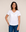 A woman with long dark hair is smiling and standing against a plain beige background, wearing The VIV Tee By You—a personalized white V-neck t-shirt from SUZI ROHER—paired with blue jeans and one hand in her pocket.