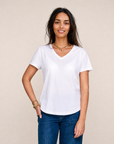 A woman with long dark hair is smiling and standing against a plain beige background, wearing The VIV Tee By You—a personalized white V-neck t-shirt from SUZI ROHER—paired with blue jeans and one hand in her pocket.