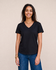 A woman with long dark hair, wearing SUZI ROHER’s The VIV Tee By You—a customizable black V-neck tee—and blue jeans, stands smiling in front of a plain beige background.
