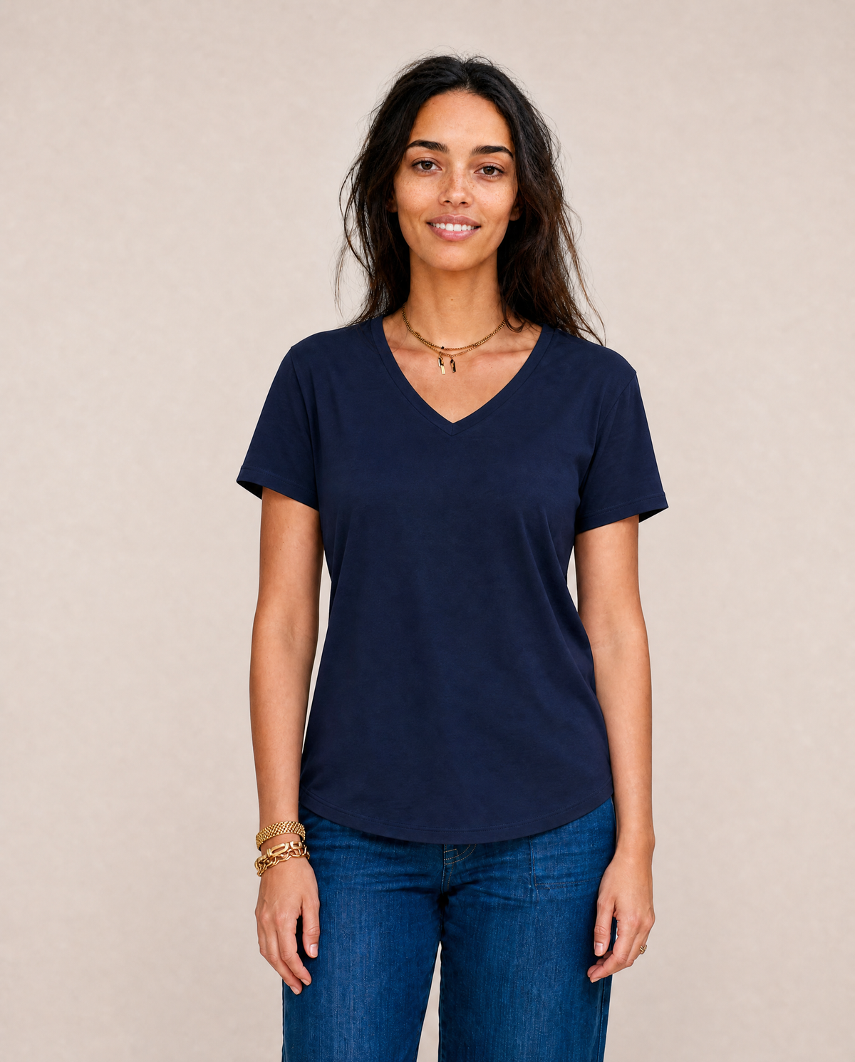 A woman with long dark hair, wearing The VIV Tee By You in navy blue from SUZI ROHER and blue jeans, stands against a neutral background, smiling gently at the camera.