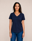 A woman with long dark hair, wearing The VIV Tee By You in navy blue from SUZI ROHER and blue jeans, stands against a neutral background, smiling gently at the camera.