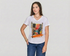 A woman with long brown hair wears The VIV Tee By You, a customizable white v-neck by SUZI ROHER featuring an abstract colorful graphic, paired with blue jeans and bracelets. She stands against a light gray background, hand on hip.